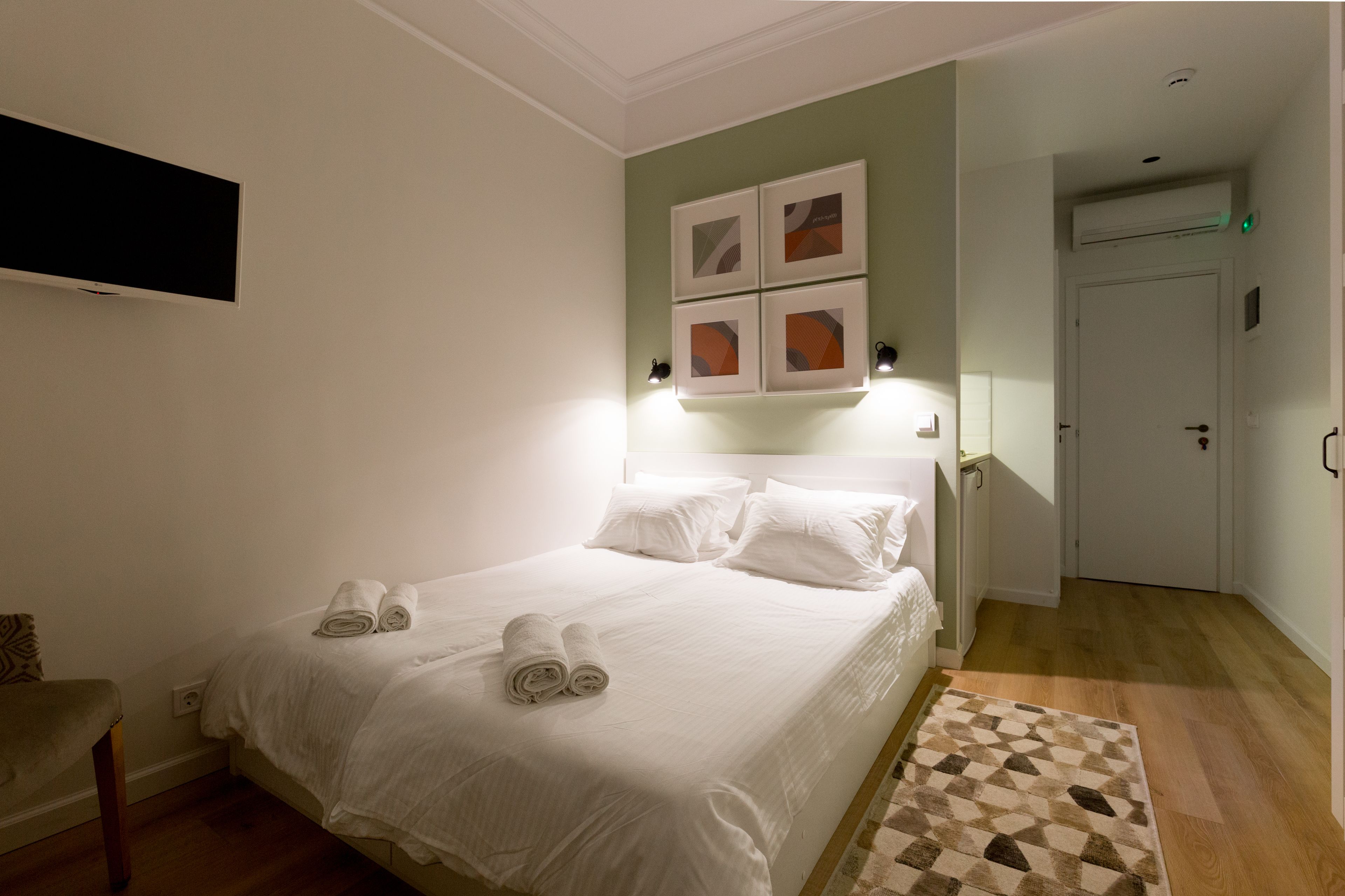 A neatly made bed with white linens and towels, a flat-screen TV mounted on the wall, and a door with a light switch.