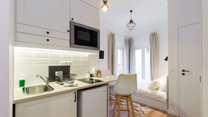 Et lille køkken med mikrobølgeovn, vask og køleskab. En barstol står ved vinduet, og en seng kan ses i det tilstødende rum.