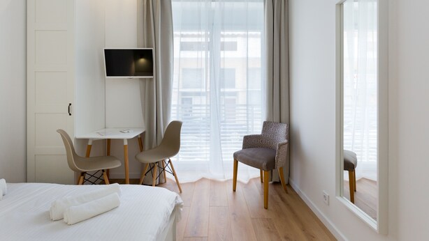 Et hotelværelse med en seng, et skrivebord, to stole, et fjernsyn og store vinduer med gardiner.