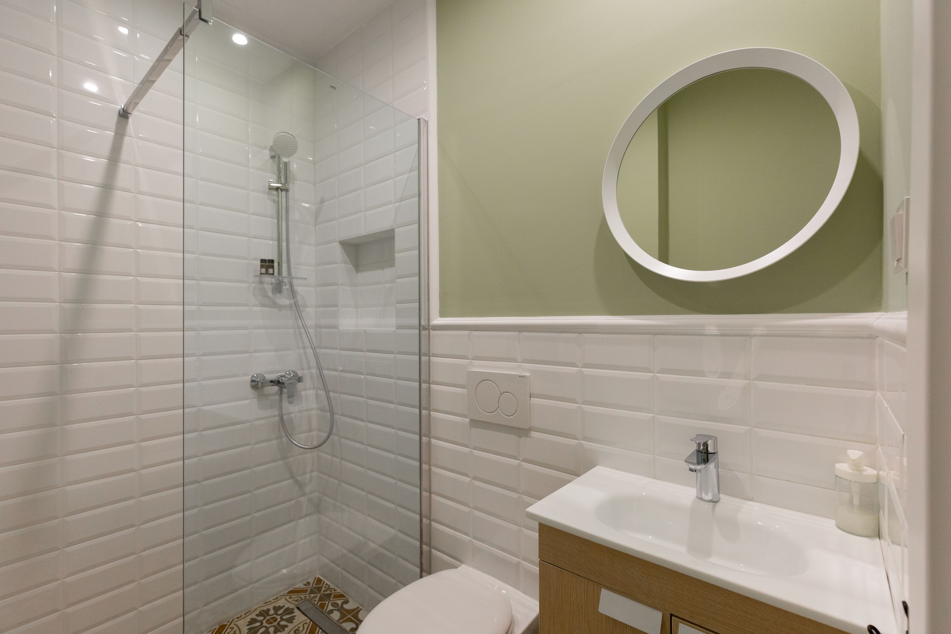 Un baño moderno con ducha de vidrio, lavamanos blanco y un espejo redondo sobre una pared verde.