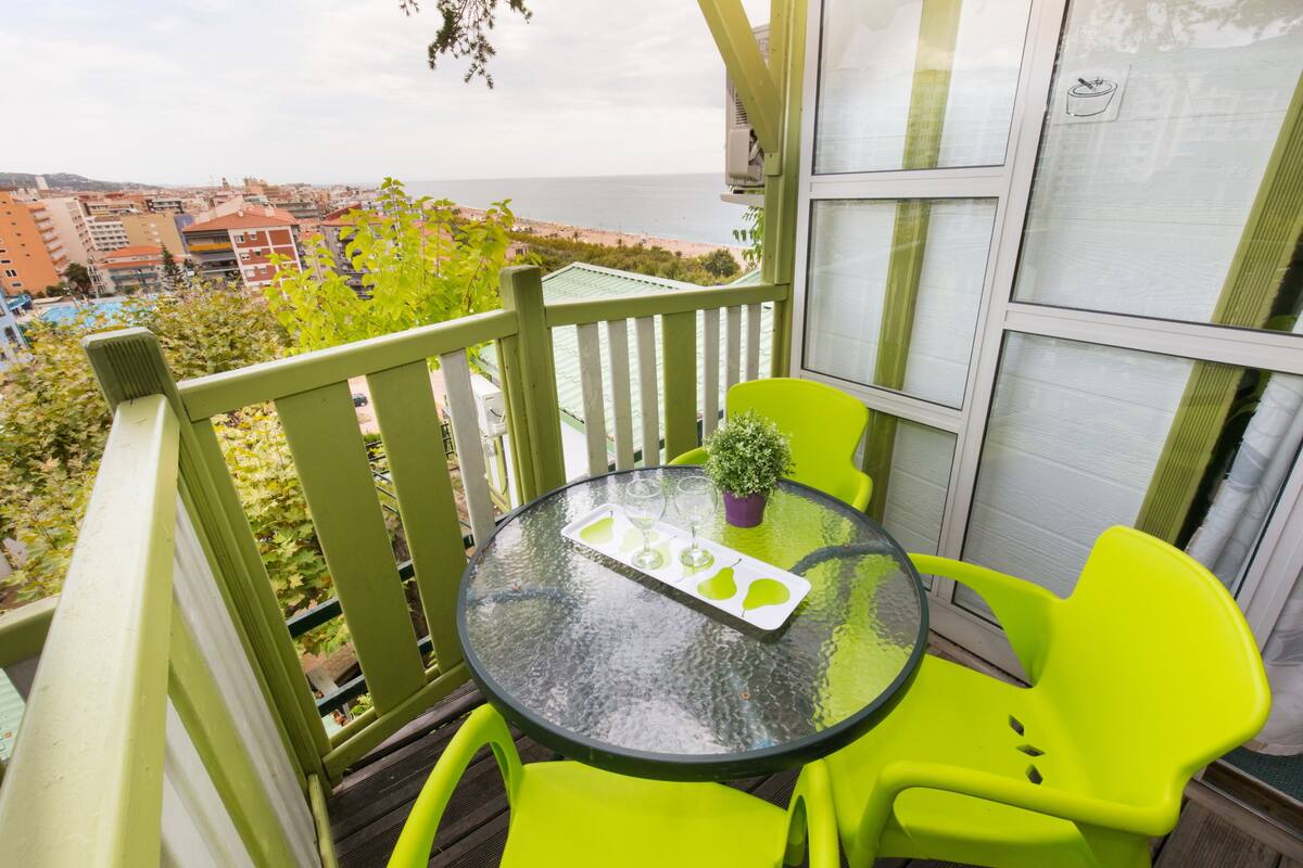 Un balcón con una mesa de vidrio y sillas de color verde brillante, con vista a una ciudad y una playa.