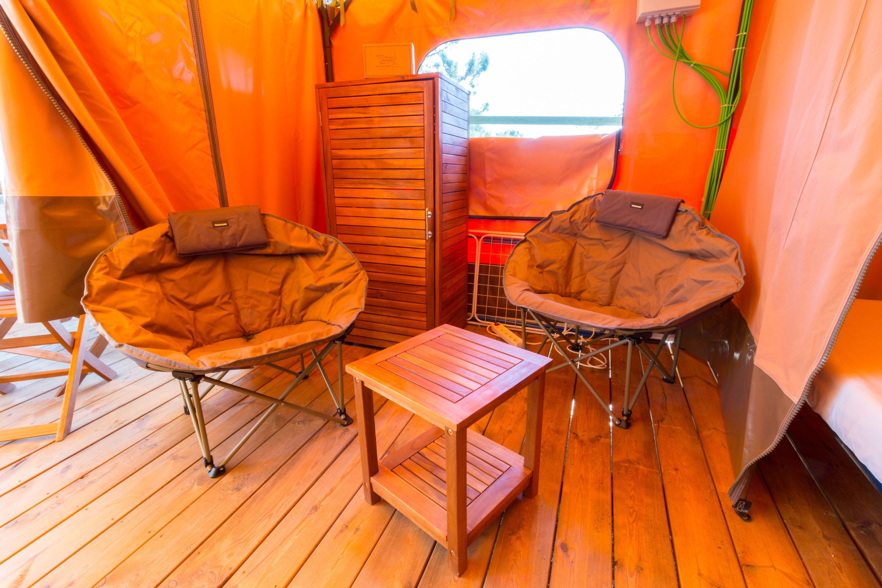 A compact, orange-themed interior with two foldable chairs, a small wooden table, and a cabinet.