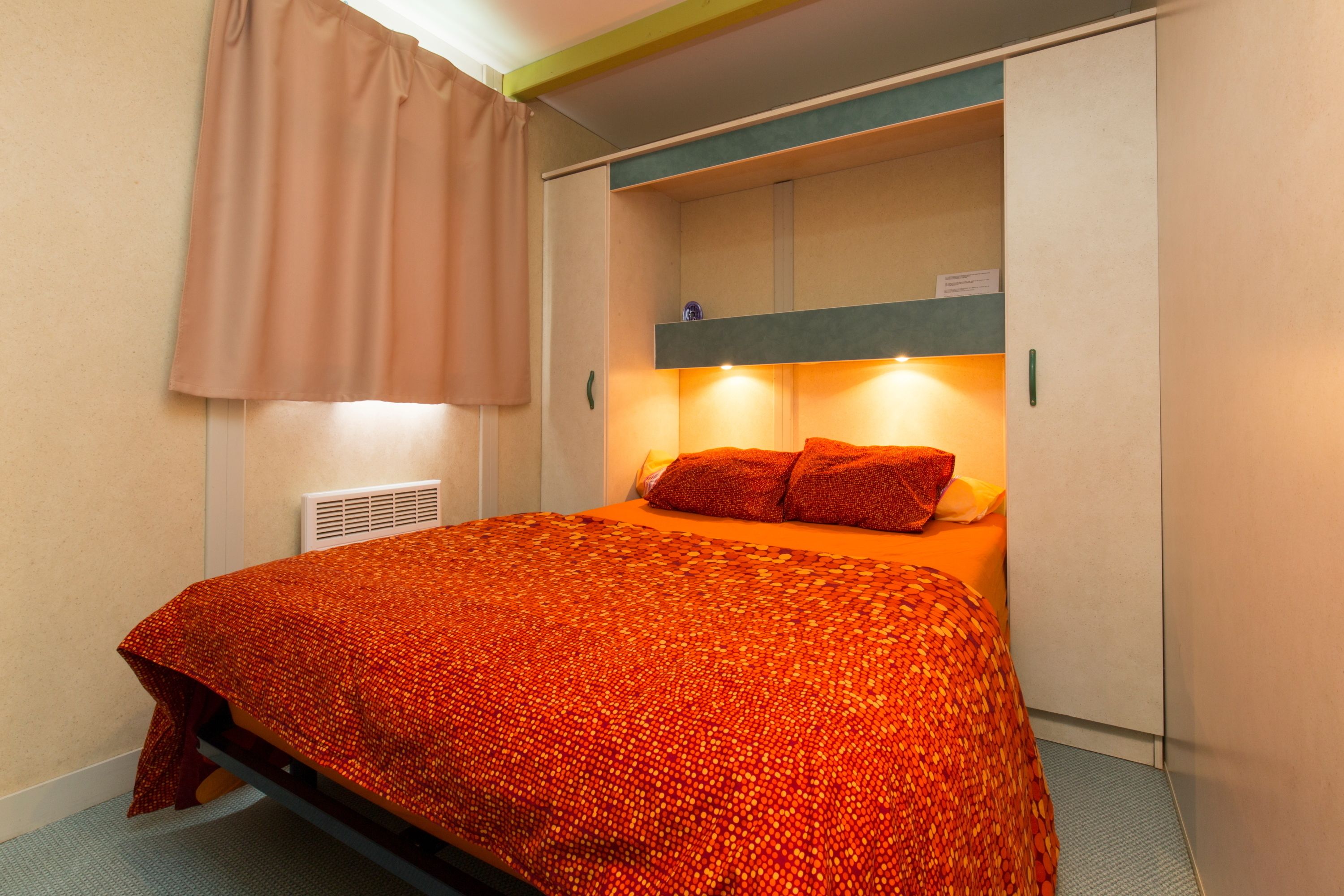 Ett kompakt hotellrum med en säng, en garderob och en liten hylla med en lampa.