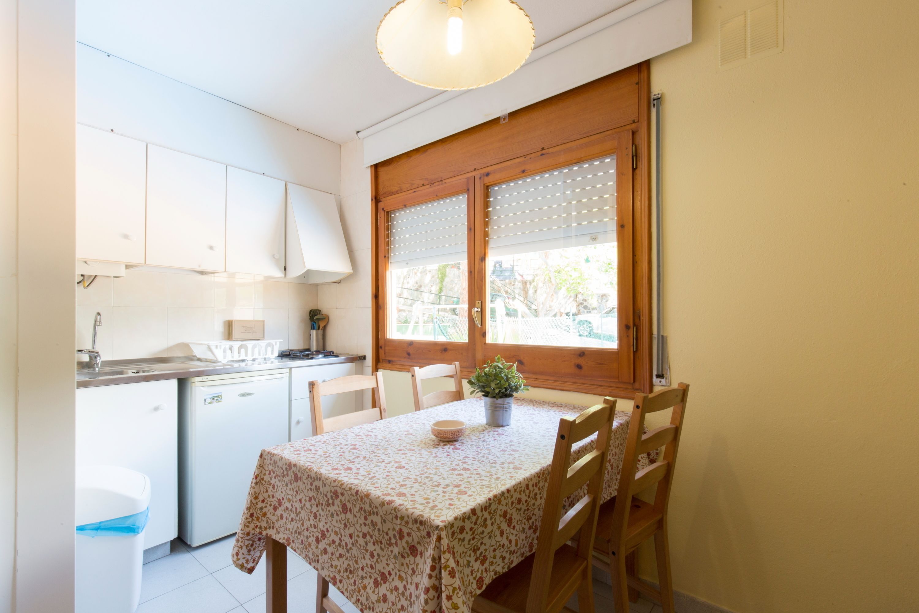 Una cocina compacta con armarios blancos, un refrigerador pequeño, una mesa de madera con sillas y una ventana con persianas.