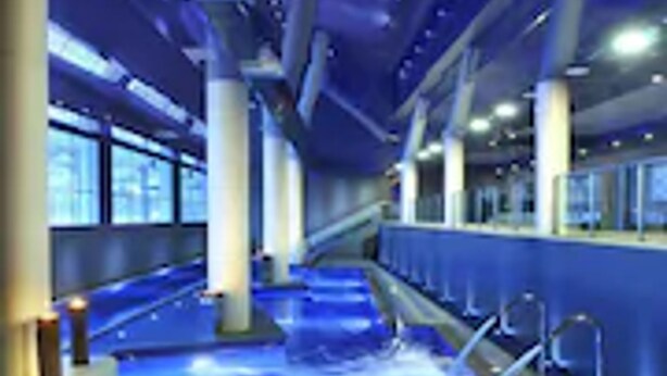 A modern indoor swimming pool with a blue-lit water feature, surrounded by columns and large windows. - Camping El Far (Calella)