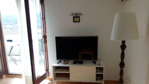 A room with a television on a white stand, a lamp, and a sliding glass door leading to an outdoor area. - Apartment Jasenka (Pag)