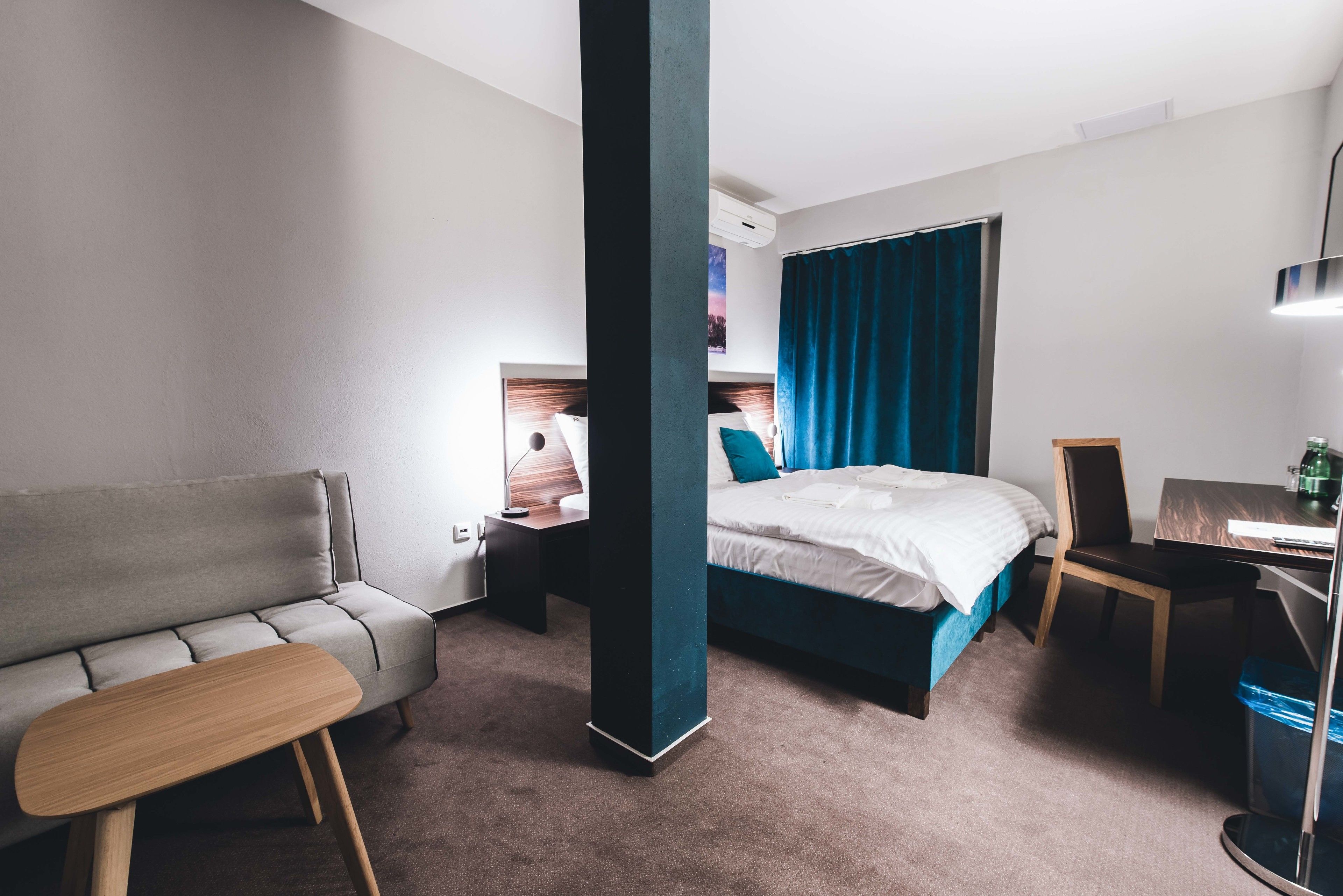 Une chambre d’hôtel moderne avec un canapé gris, un lit avec des rideaux bleus, un bureau en bois et une chaise.