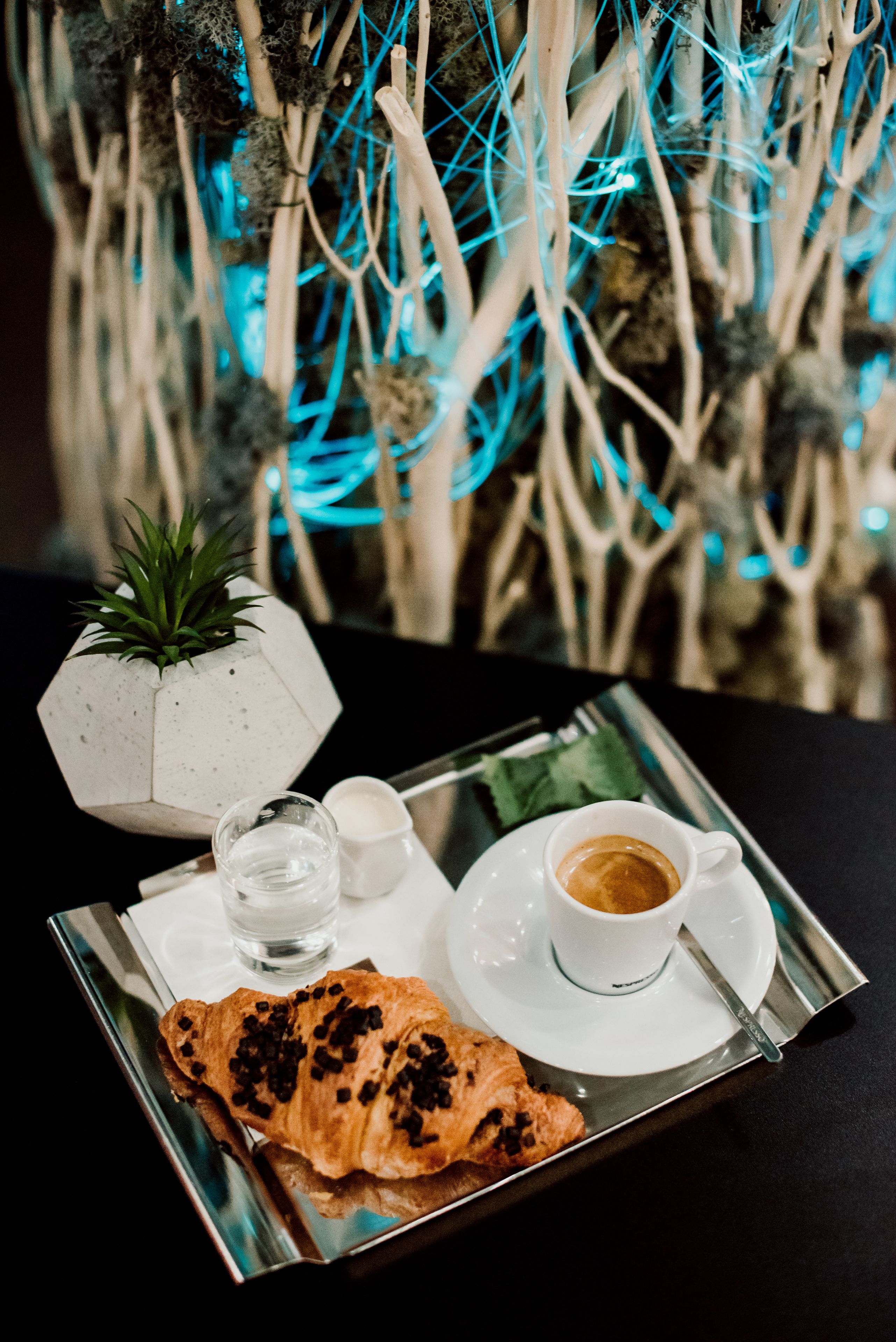 Uma bandeja com um croissant, um copo de água, um copo de café e uma pequena tigela de açúcar sobre uma mesa, com um painel decorativo ao fundo.