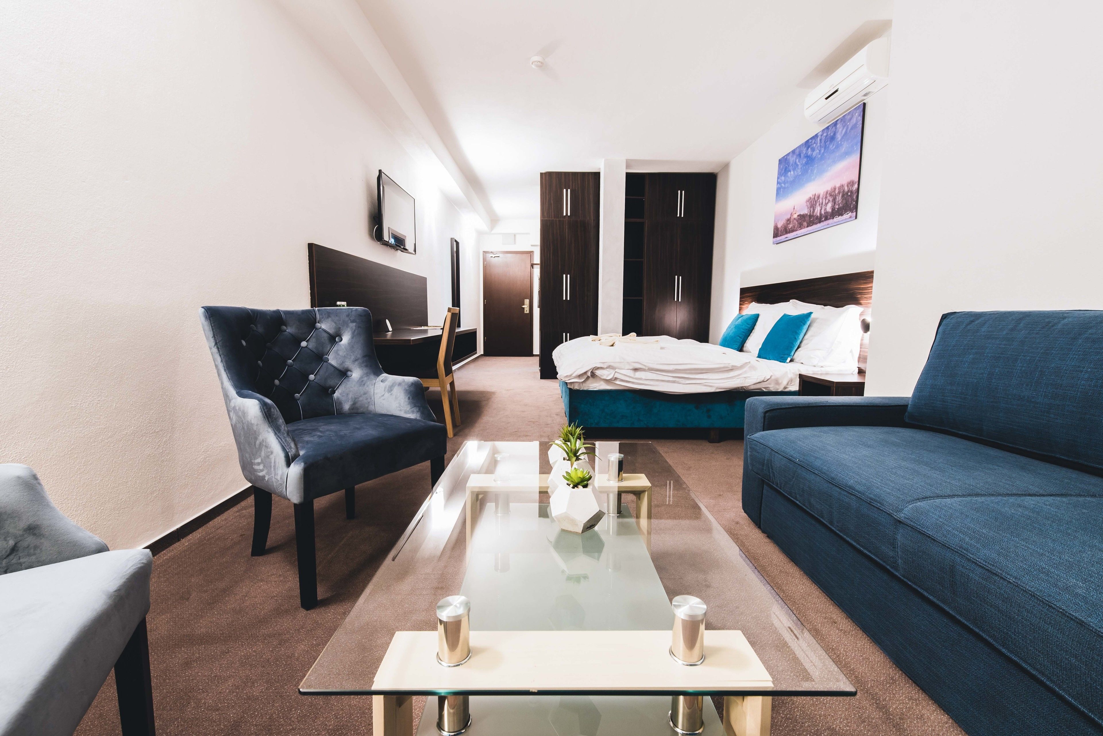 Una habitación de hotel moderna con una mesa de centro de vidrio, un sofá azul, un sillón oscuro y una cama con ropa de cama azul.