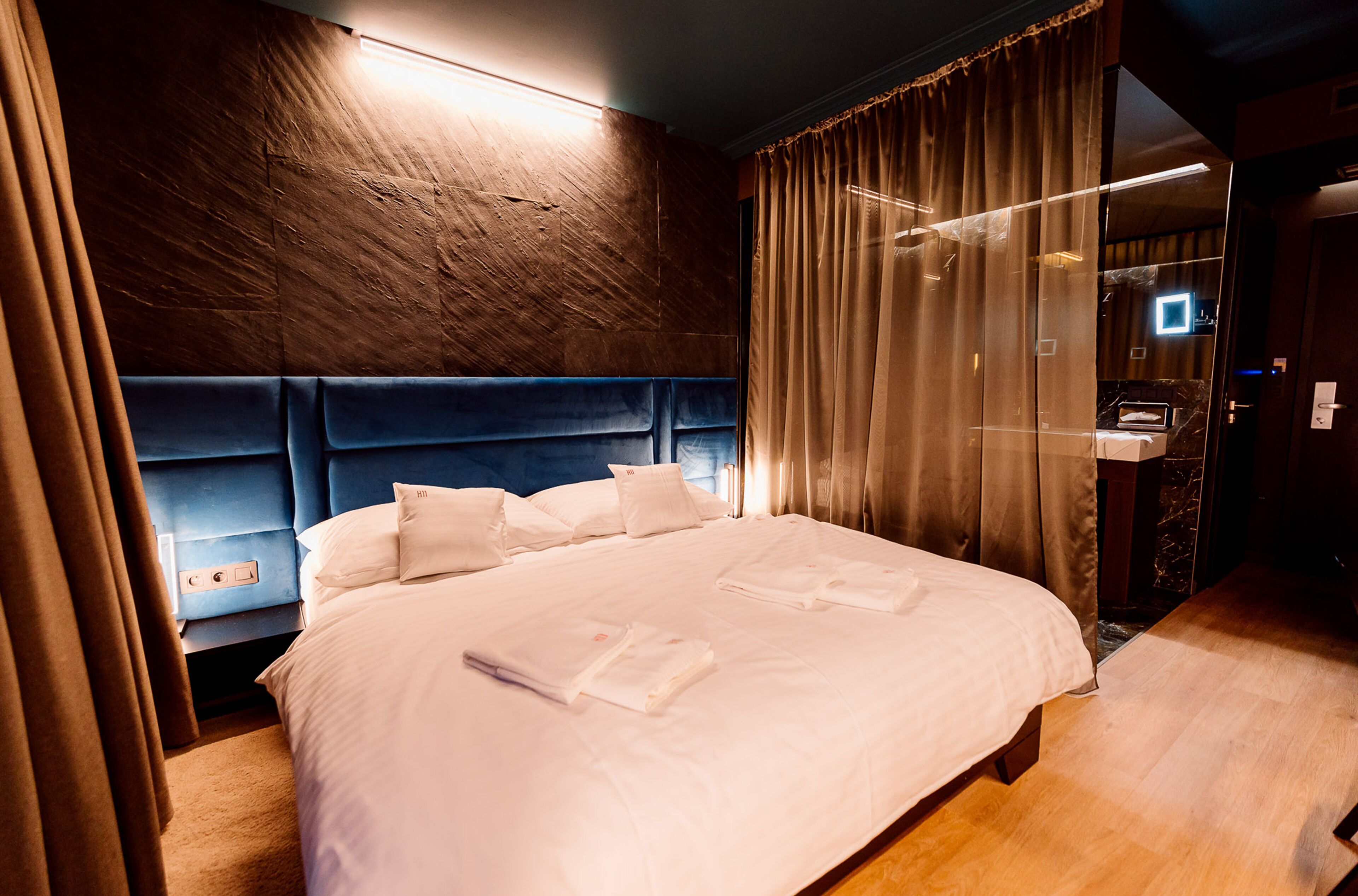 Un lit bien fait, avec du linge de lit et des serviettes blanches, une tête de lit en bois et une salle de bain moderne dotée d’une douche en verre.