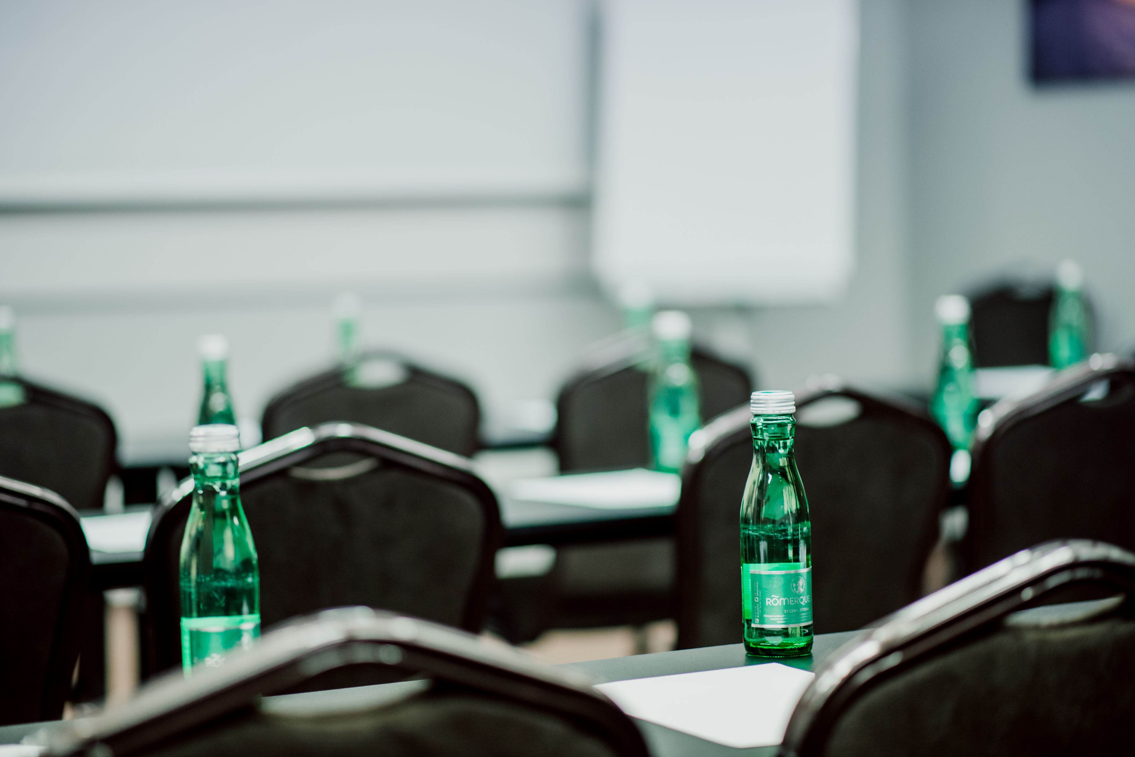 A conference room with rows of black chairs and tables, each with a green bottle of water.
