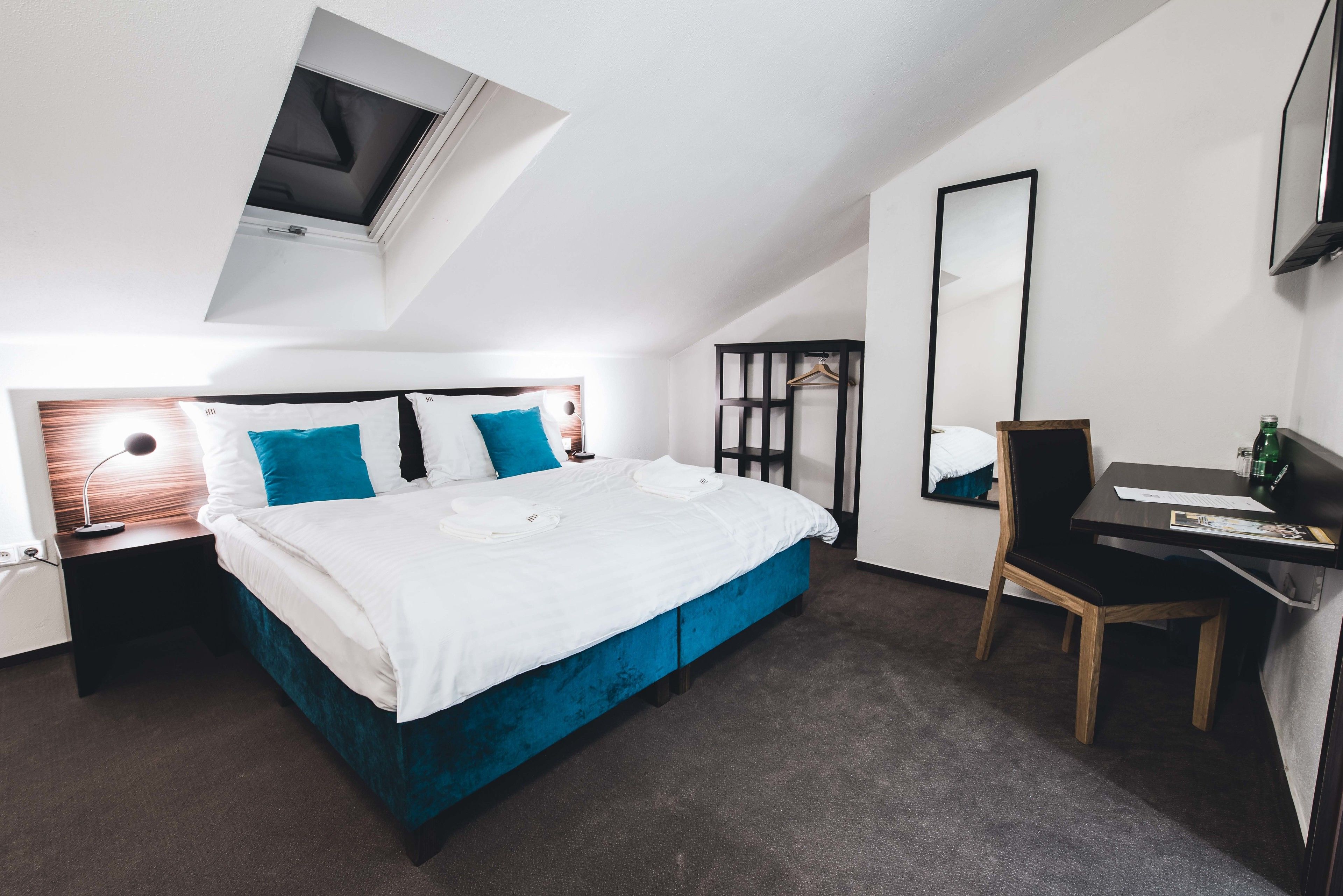 A modern hotel room with a large bed, a desk, a chair, a mirror, and a skylight.