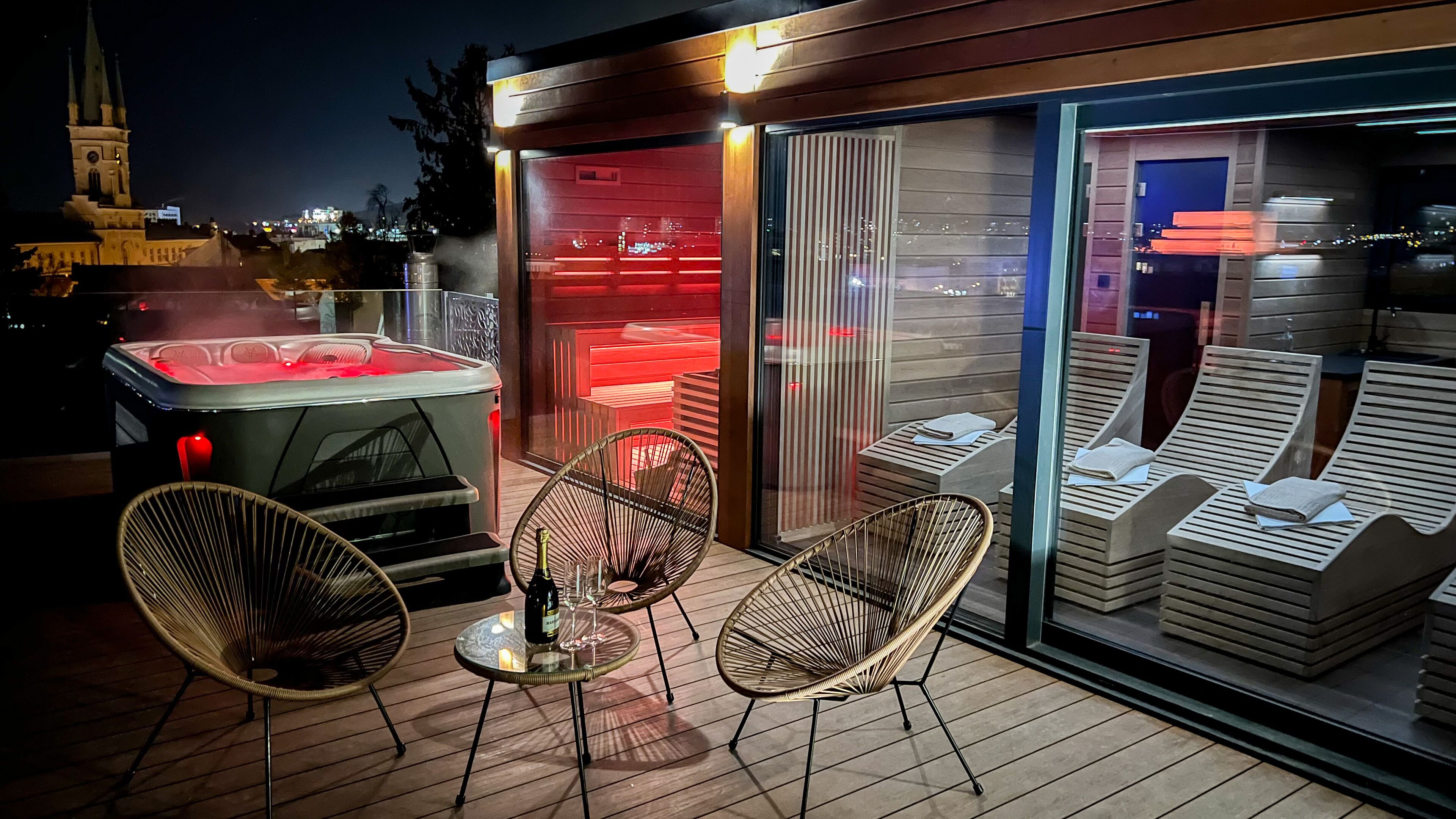 Un bain à remous éclairé d’une lumière rouge, deux fauteuils en osier et des chaises longues avec des serviettes.