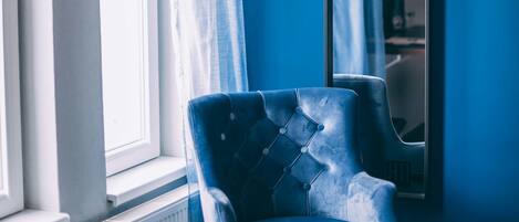 Ein Zimmer mit großem Fenster, blauem Sessel und Spiegel an der Wand.