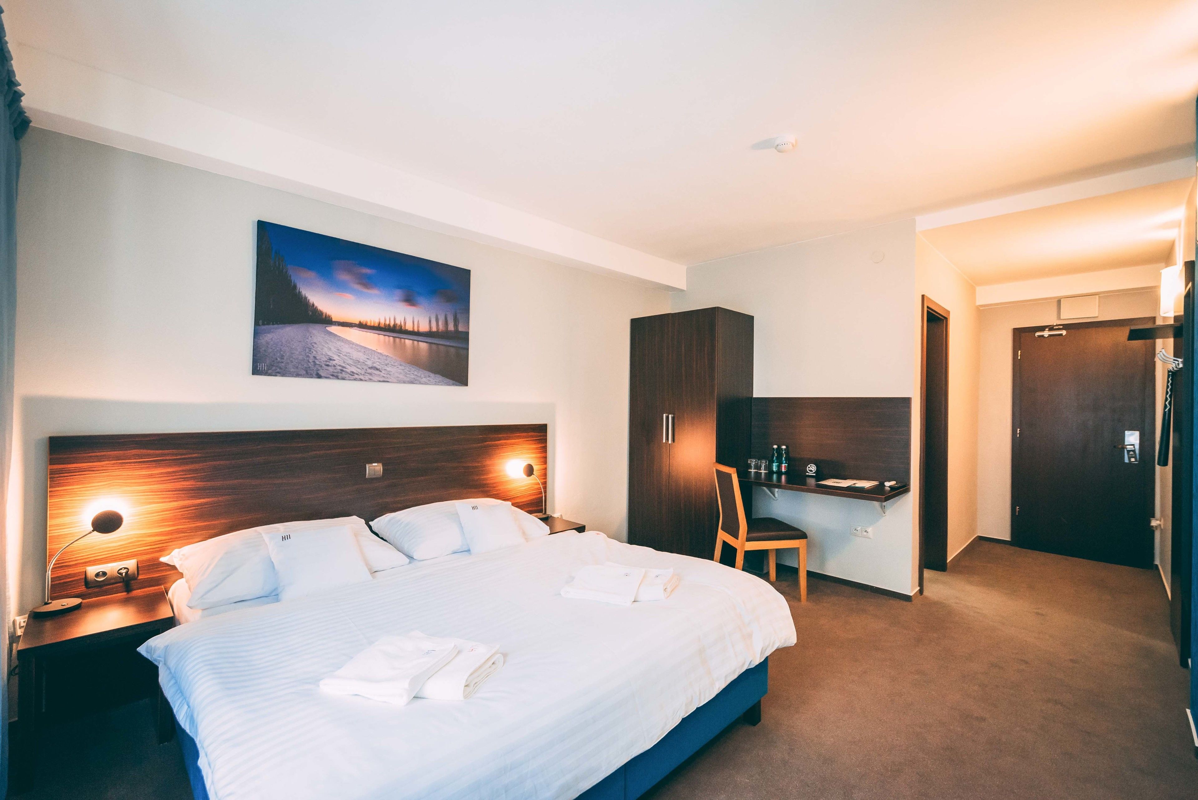A hotel room with a large bed, a desk, a chair, a wardrobe, and a painting on the wall.
