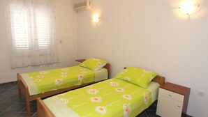 A room with two single beds, a small bedside table, a wall-mounted air conditioner, and a window with curtains.