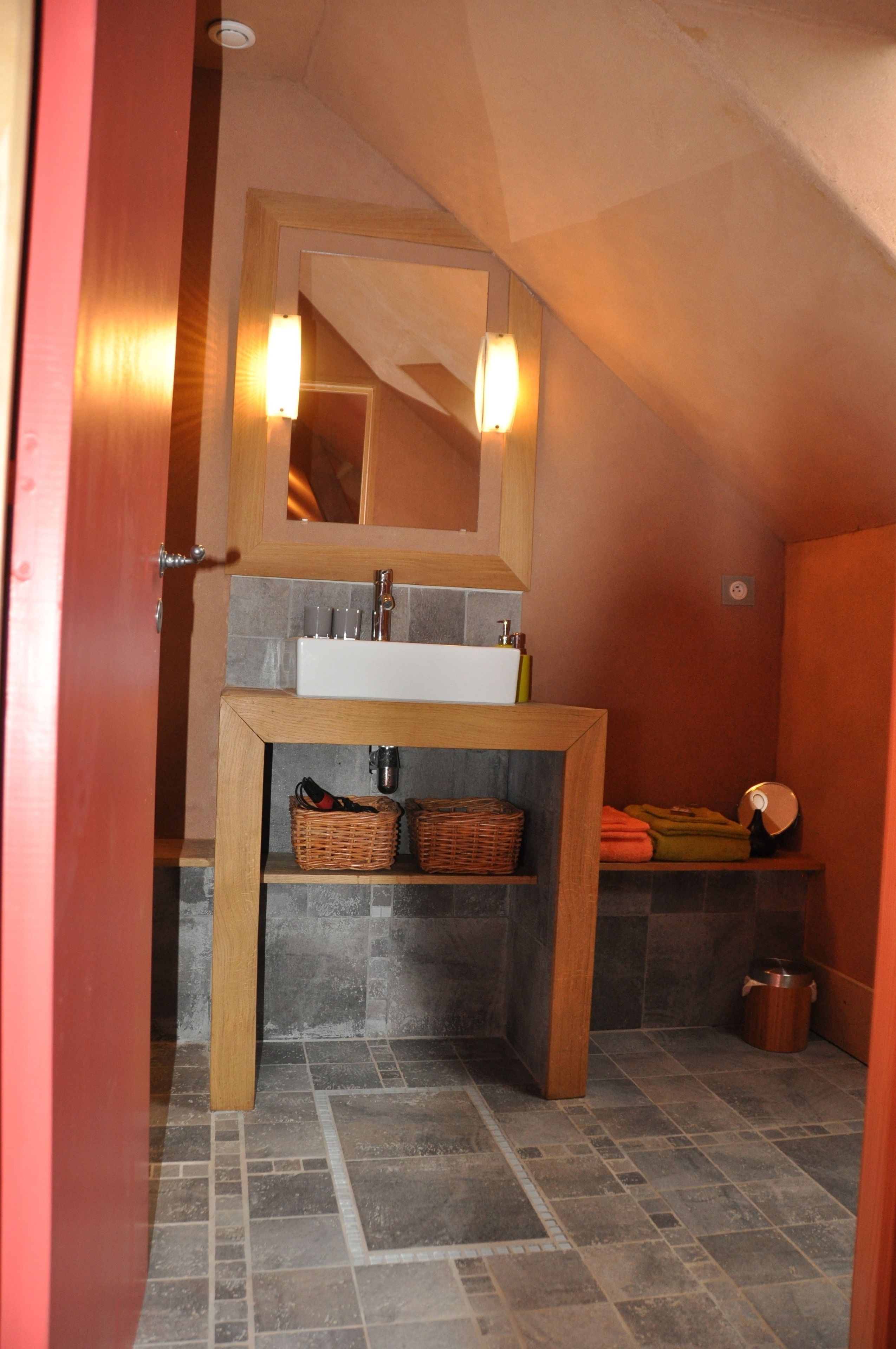 Une salle de bain avec un meuble-lavabo en bois, un lavabo blanc et un miroir au-dessus. On y trouve deux paniers en osier sous le meuble-lavabo et un porte-serviettes avec des serviettes.