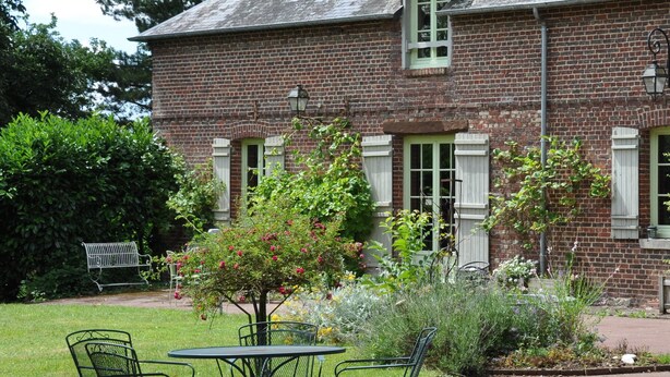 A brick building with a garden area featuring a table and chairs, a bench, and various plants. - Les Jardins de Beauve (Héricourt-sur-Thérain)