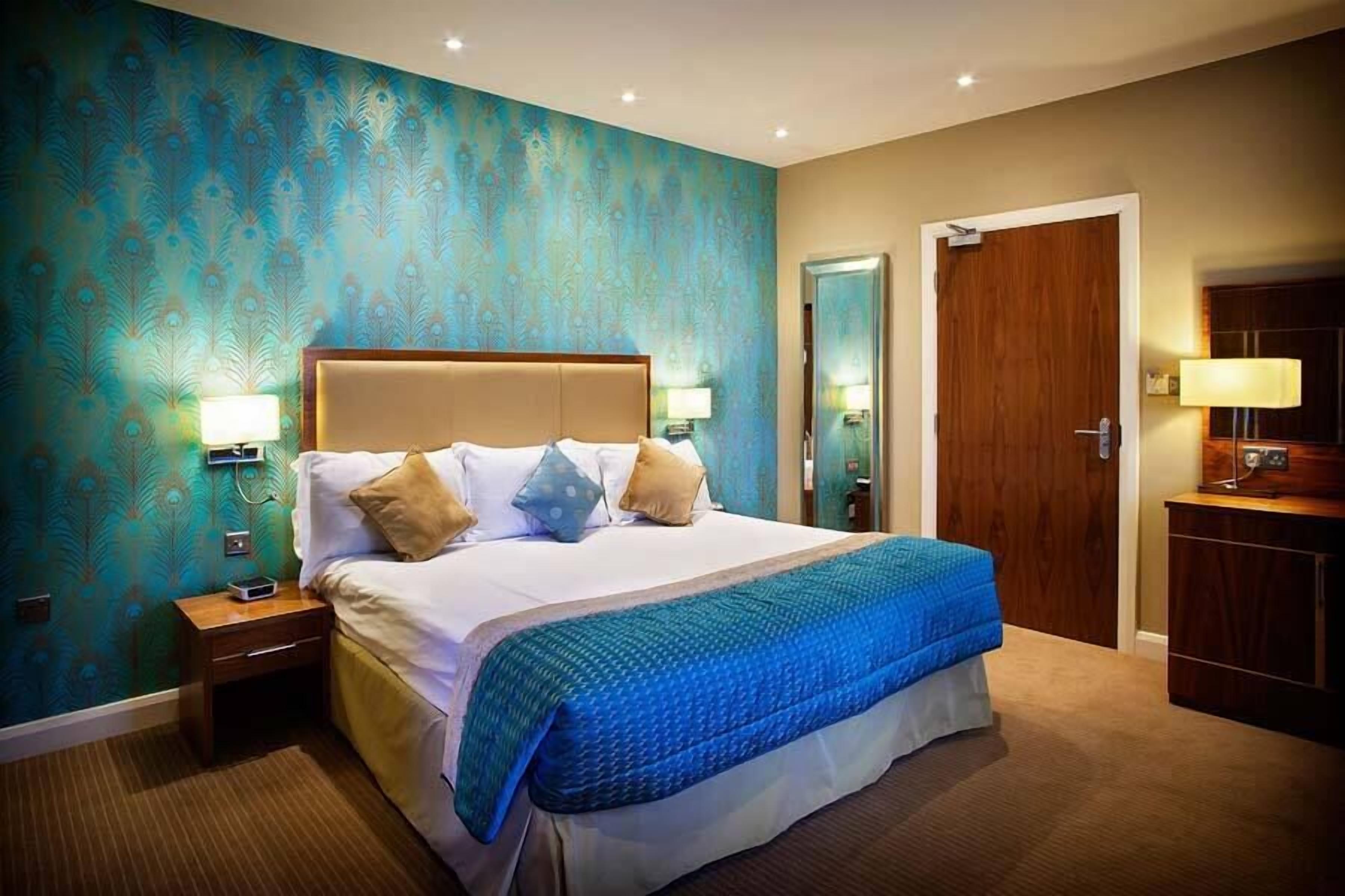 A hotel room with a large bed, two bedside tables with lamps, a wooden wardrobe, and a teal patterned wall.