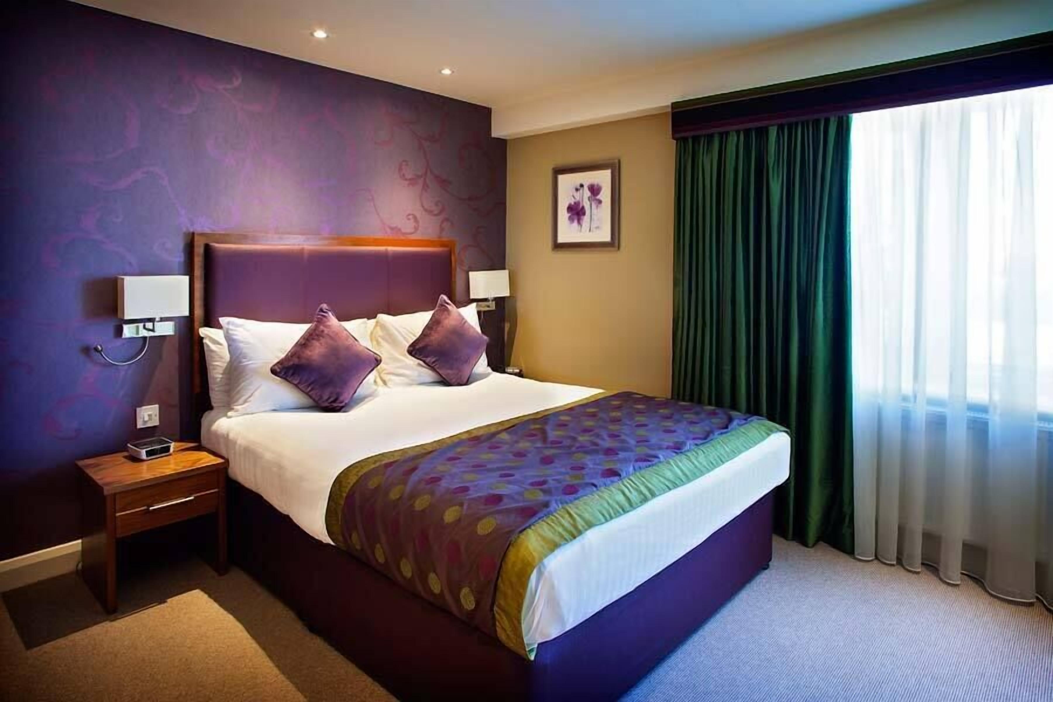 A hotel room with a large bed, two bedside tables, a nightstand, a framed picture on the wall, and a window with curtains.