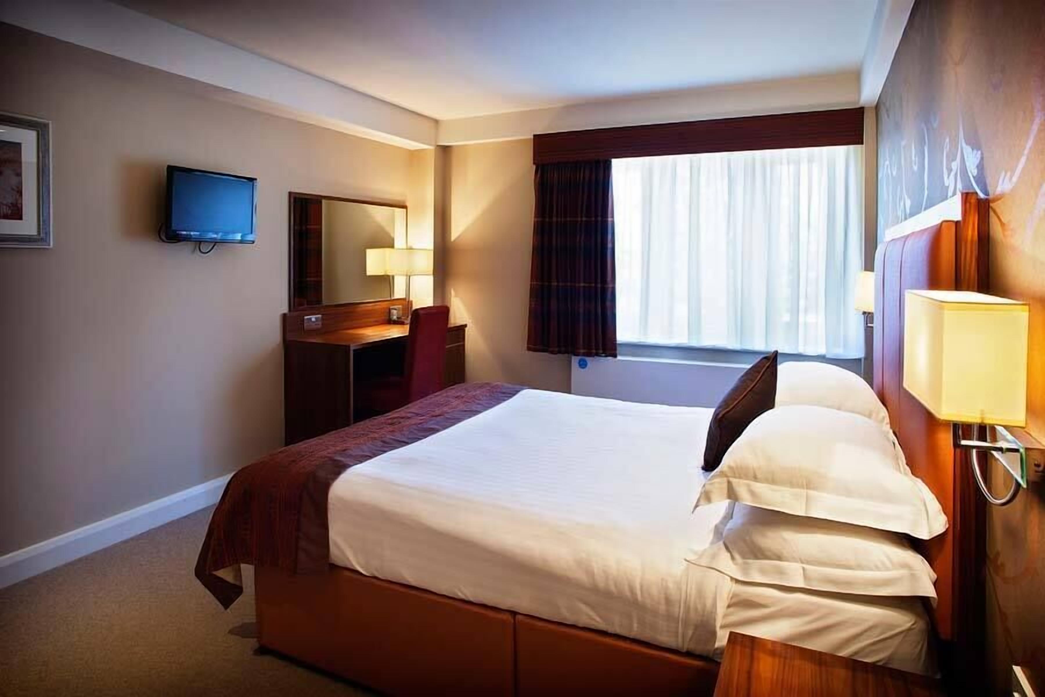 A hotel room with a large bed, a desk with a chair, a television, a window with curtains, and a wall-mounted lamp.