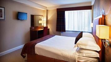 A hotel room with a large bed, a desk with a chair, a television, a window with curtains, and a wall-mounted lamp.