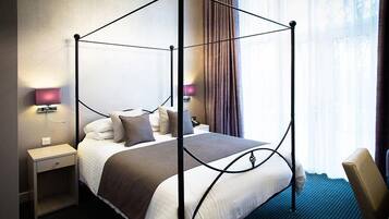 A four-poster bed with a dark metal frame, white bedding, and a brown throw blanket. There is a nightstand with a lamp and a chair to the right.