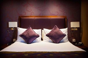 Habitación de hotel con una cama grande, dos almohadas moradas, un cabecero de madera y lámparas de noche.