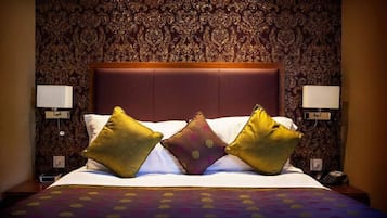 Una habitación de hotel con papel tapiz estampado, una cama grande con cabecera, dos lámparas de noche y un teléfono en la mesita.