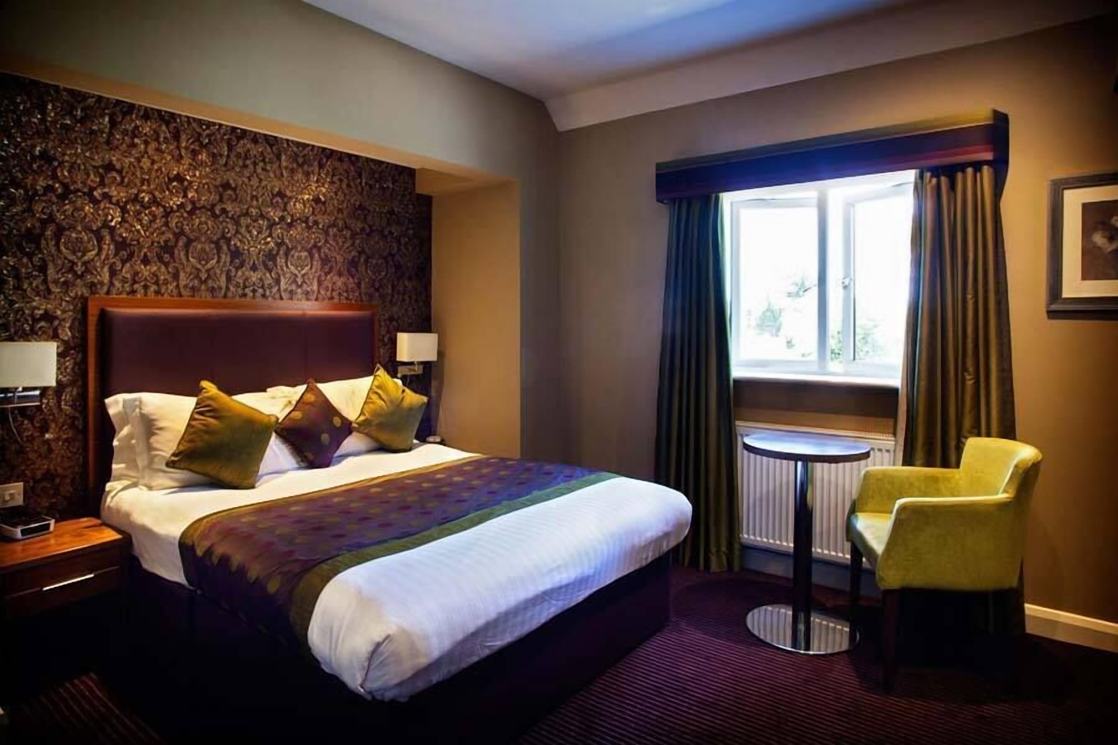A hotel room with a large bed, a small round table, a yellow armchair, and a window with curtains.