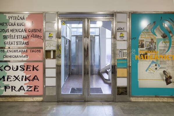 Um corredor com porta de vidro que dá para um lance de escadas. Há placas nas paredes anunciando cozinha mexicana e um estabelecimento chamado 'Kousek Mexika Praze'.
