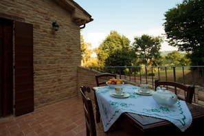Une table dressée à l’extérieur, avec une théière, des tasses et un saladier rempli de fruits, le tout posé sur une nappe.