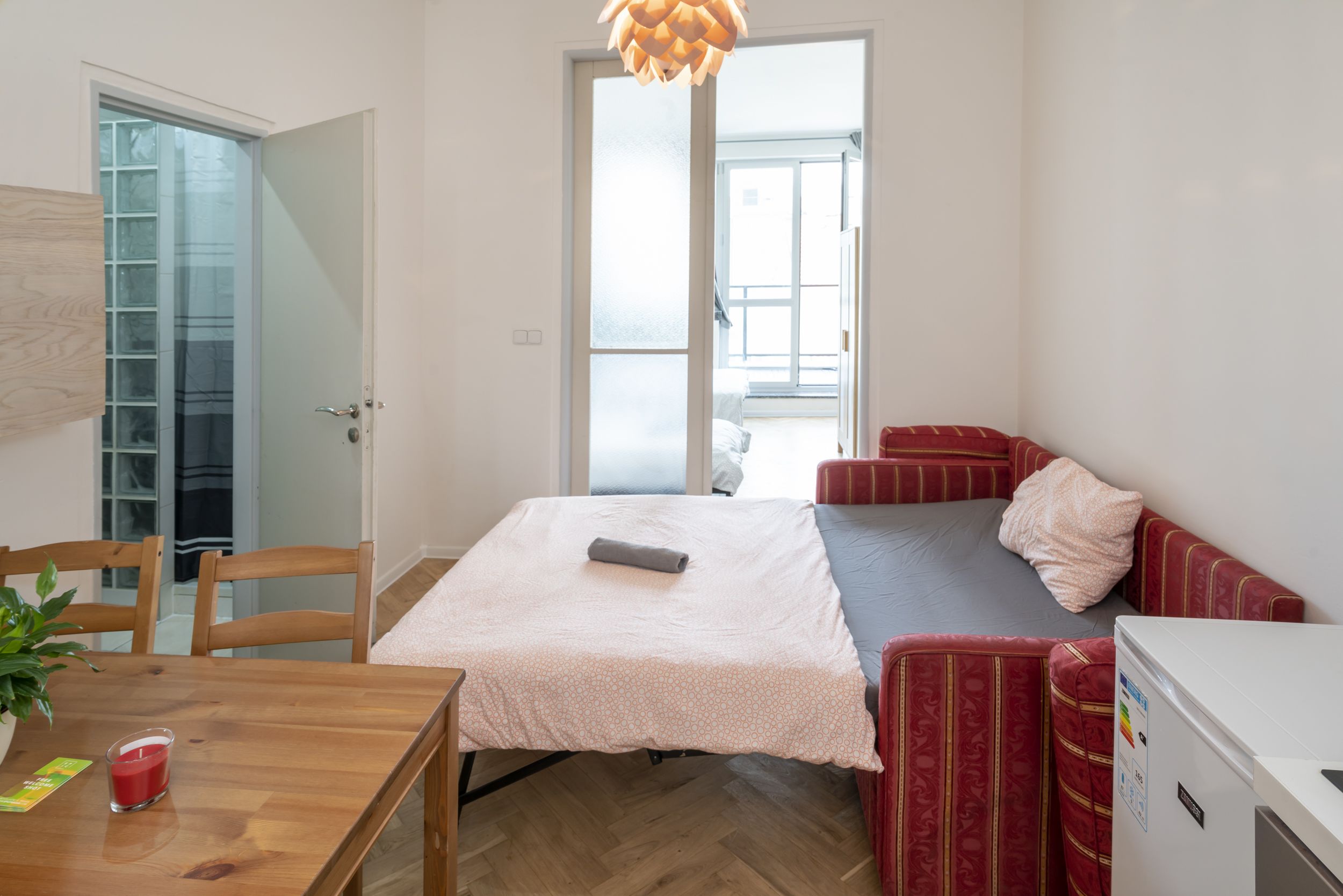 Une pièce de taille modeste comprenant un lit, une petite table avec une plante, un canapé rouge et un réfrigérateur blanc.