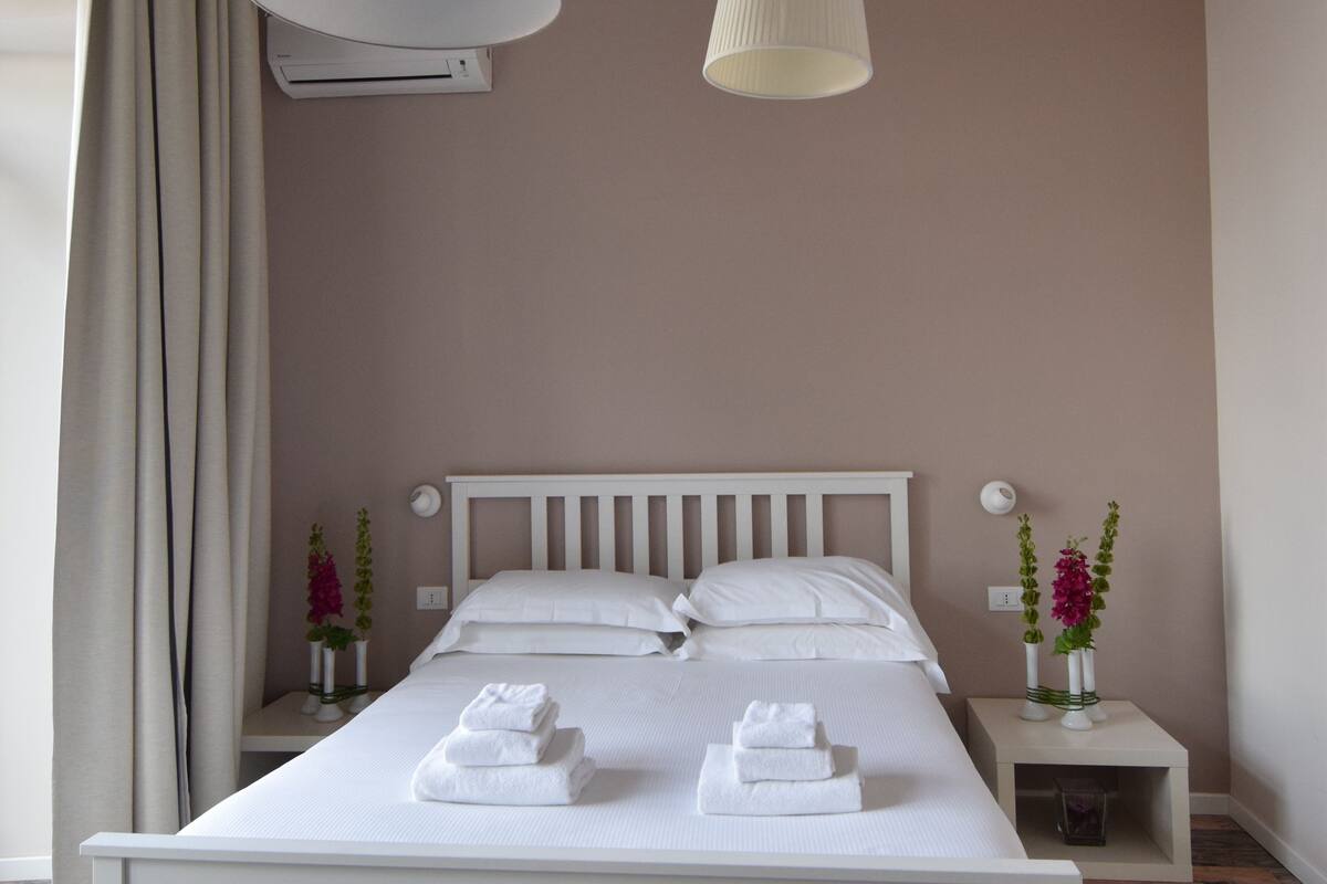 Un lit bien fait, avec du linge de lit et des serviettes blanches, une table de chevet blanche ornée d’un vase de fleurs et une grande fenêtre avec des rideaux.