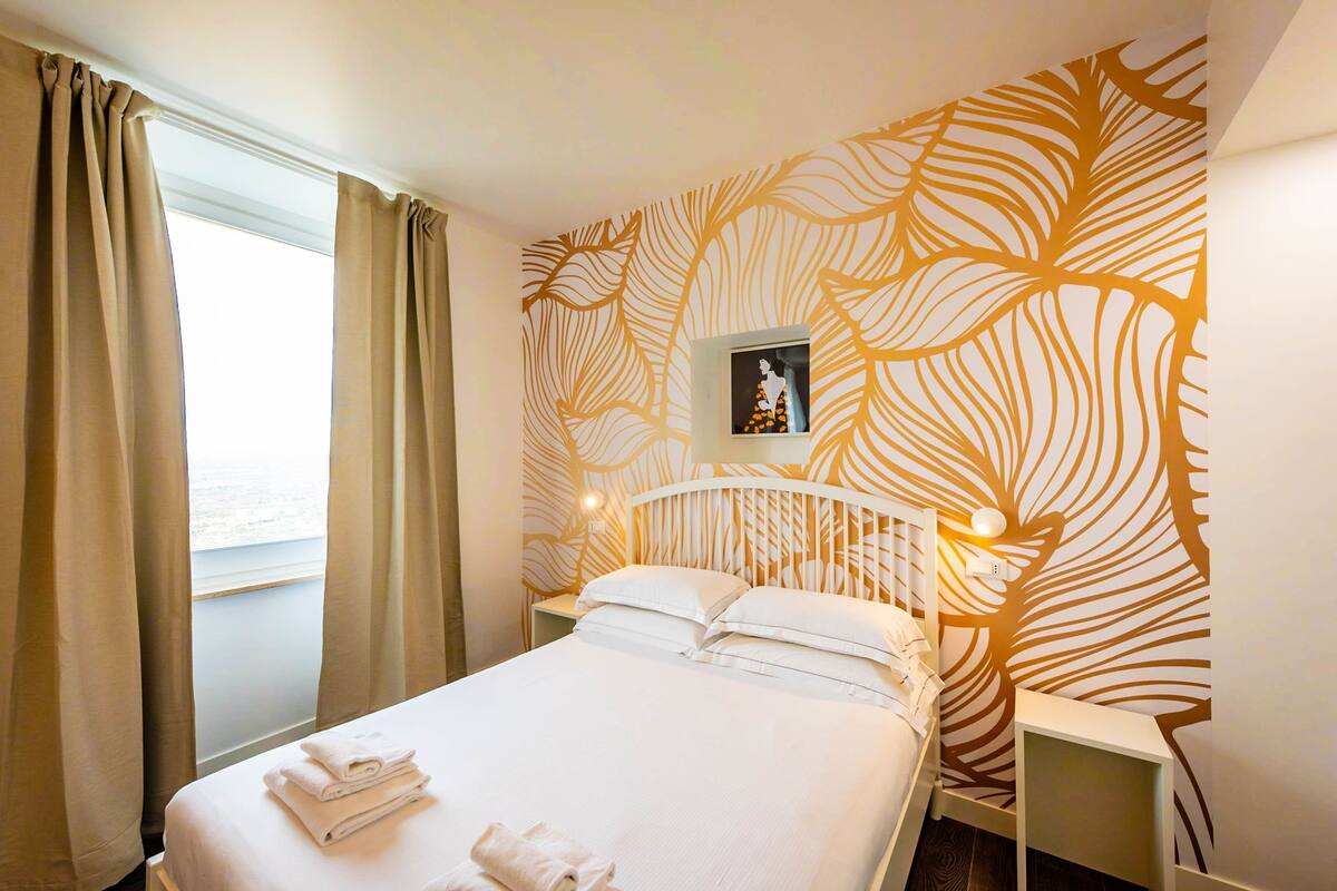 Une chambre d’hôtel avec un lit, des rideaux, une table de chevet, un mur orné d’un motif de feuilles et un tableau accroché au mur.