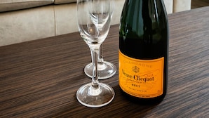 Uma garrafa de champanhe Veuve Clicquot e dois copos de vinho vazios sobre uma mesa de madeira numa sala de estar com um sofá bege e um painel decorativo ao fundo.