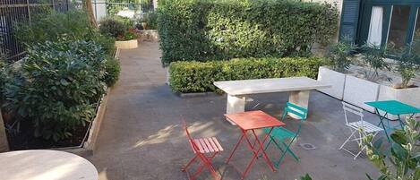 A courtyard with a concrete table, two red folding chairs, a green folding chair, and a small white table. There are potted plants and a hedge in the background.