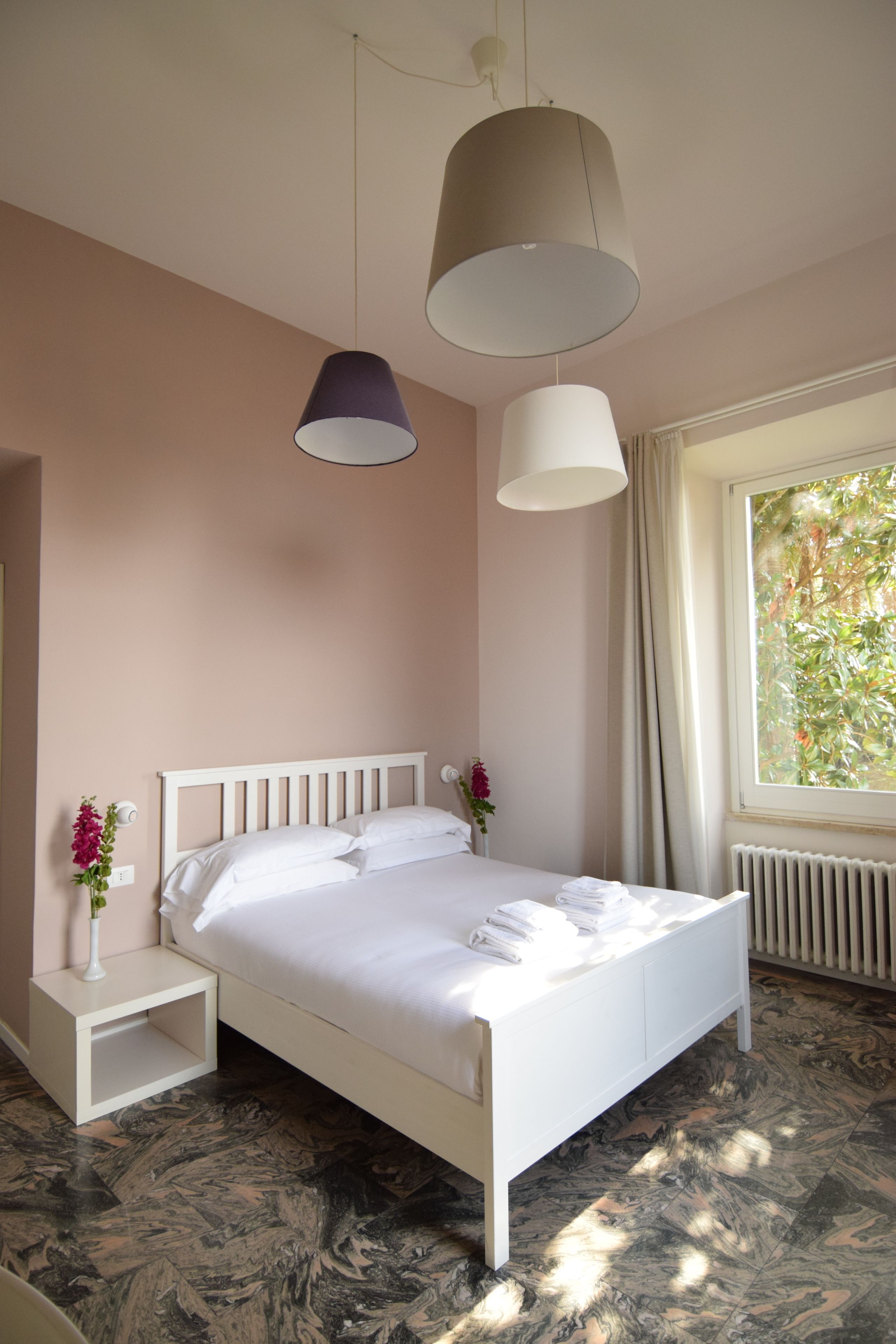 Ein Schlafzimmer mit einem weißen Bett, zwei Nachttischen, einem Fenster mit Blick ins Grüne und einer Heizung.