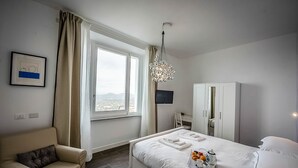 Quarto de hotel com cama, sofá, guarda-roupa, televisão e janela com cortinas.