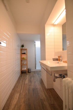Un baño con paredes de azulejos blancos, un estante de madera, un lavabo blanco con espejo y una zona de ducha.
