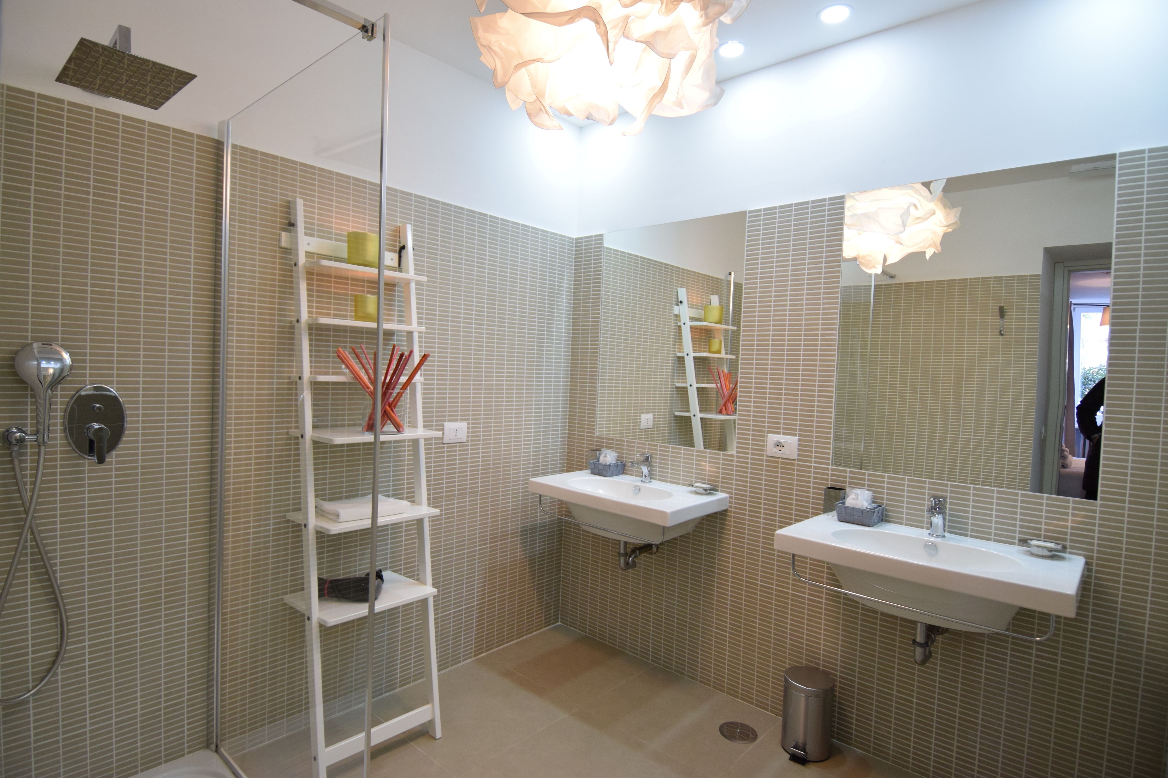Un baño moderno con dos lavamanos, ducha y zona de ducha con cerramiento de vidrio. Hay estantes con artículos de aseo y un cubo de basura.