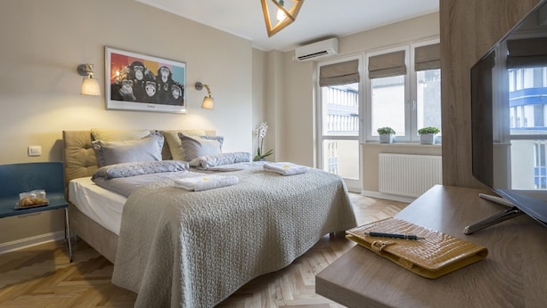A bedroom with a bed, bedside table, lamp, air conditioning unit, window with view, and a TV.