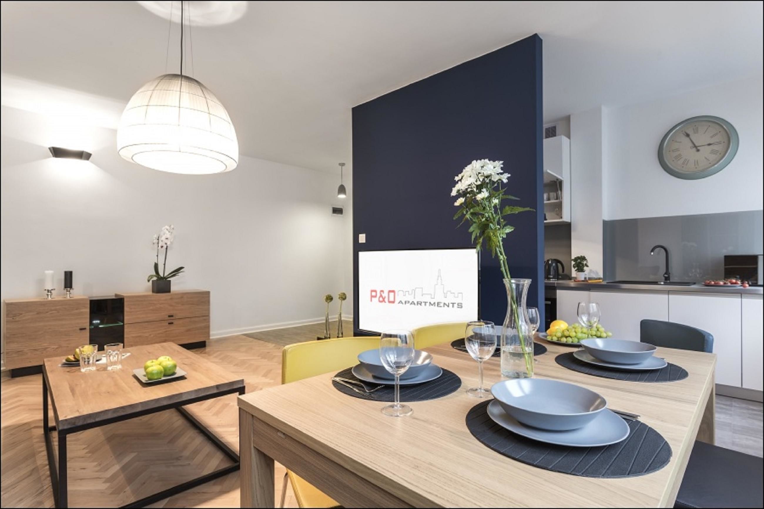 Un espace repas moderne avec une table en bois dressée pour quatre personnes, un vase rempli de fleurs et une enseigne indiquant « P&O Apartments ».