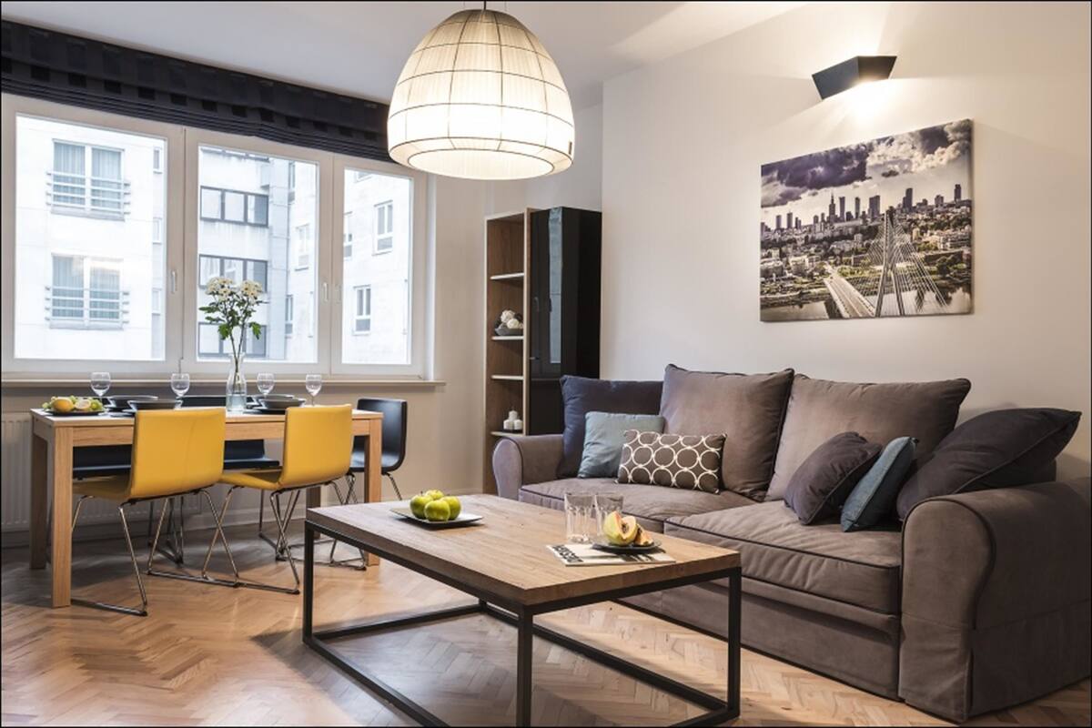 Una sala de estar moderna con un sofá, una mesa de comedor con sillas, una lámpara colgante y un cuadro de un paisaje urbano en la pared.