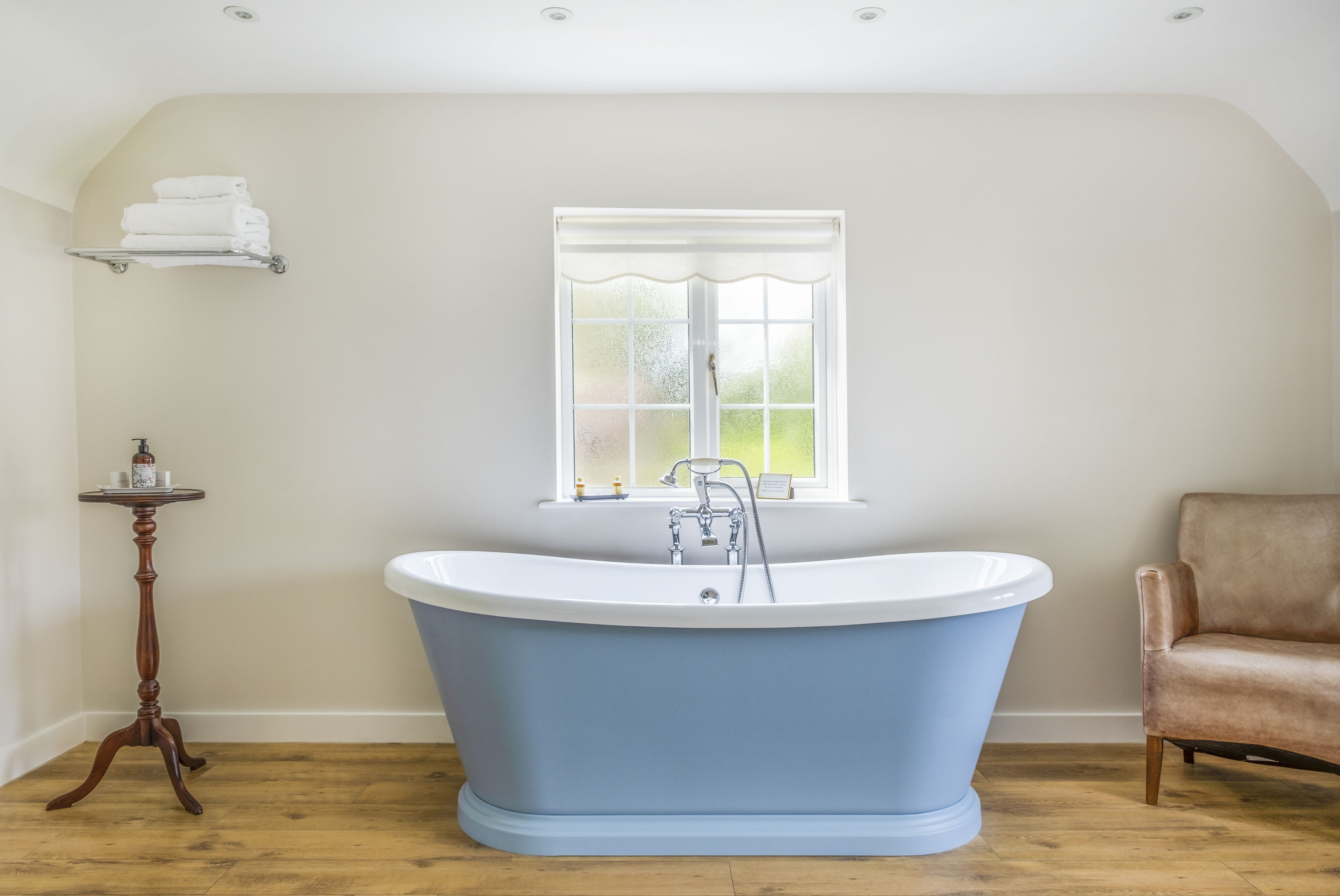 Une salle de bain avec une baignoire sur pieds, une fenêtre à verre dépoli, un plancher en bois et un fauteuil en cuir marron.