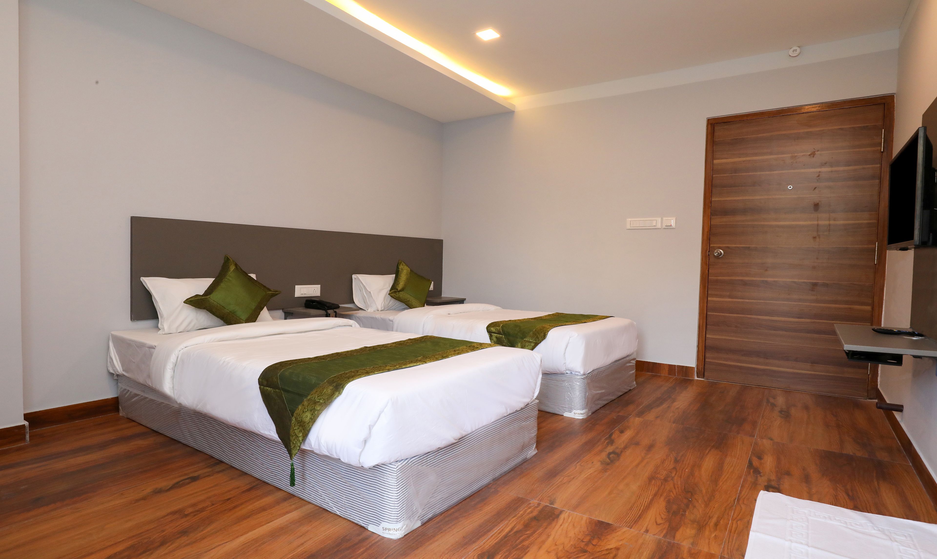 Een hotelkamer met twee bedden, een houten vloer, een grote houten deur en een flatscreen televisie aan de muur.