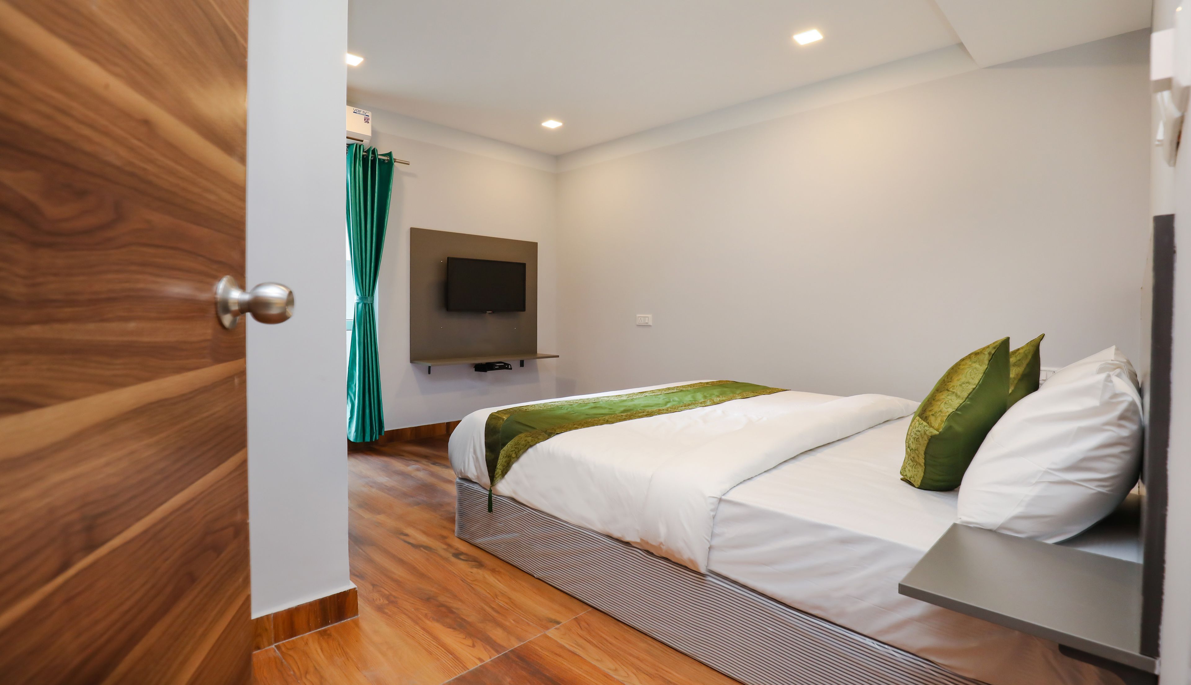 A modern hotel room with a wooden floor, a bed with white bedding and green pillows, a flat-screen TV mounted on the wall, and a teal curtain.