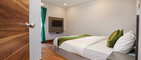 A modern hotel room with a wooden floor, a bed with white bedding and green pillows, a flat-screen TV mounted on the wall, and a teal curtain.