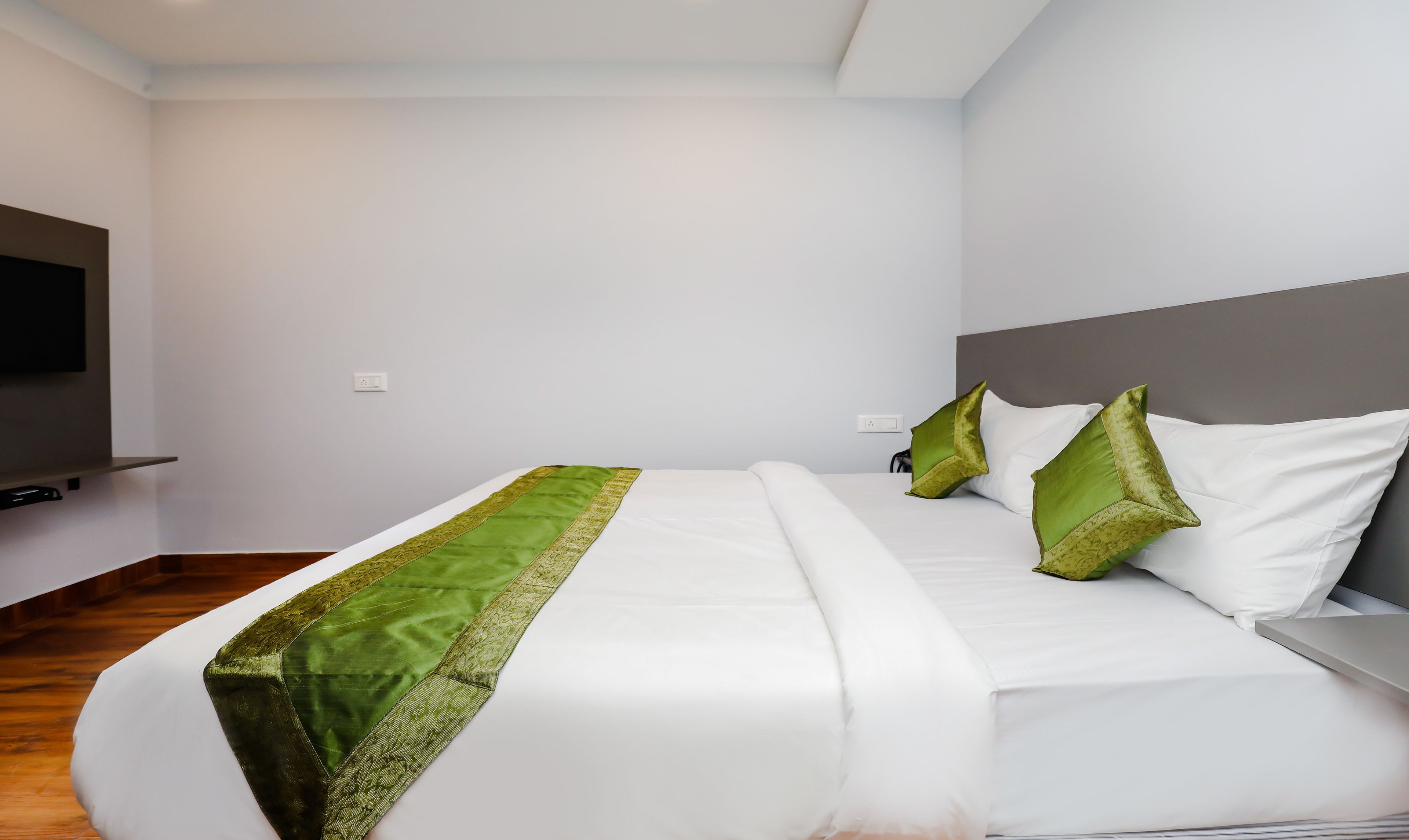 Una habitación de hotel moderna con una cama grande, almohadones decorativos verdes y un televisor de pantalla plana montado en la pared.