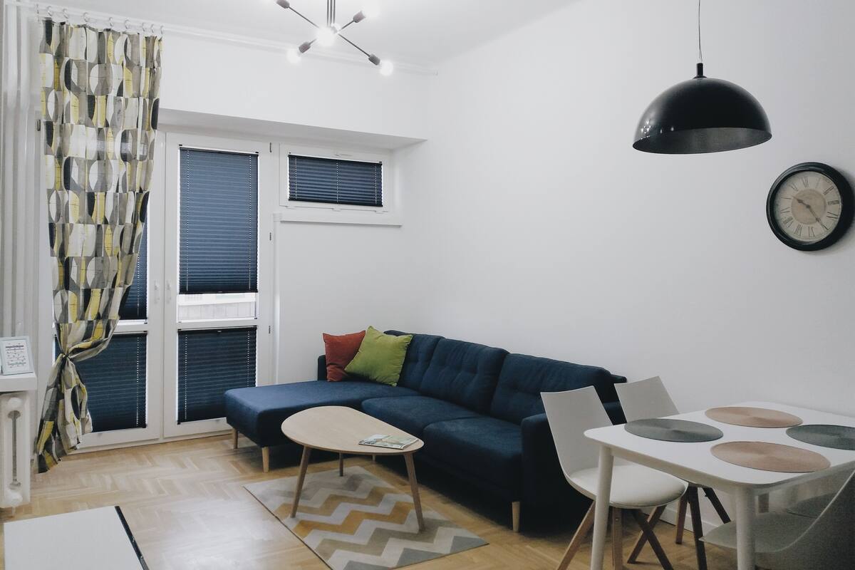 A modern living room with a blue sofa, a small table, and a dining area with chairs.